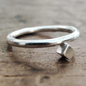 May include: A silver ring featuring a small, square, geometric detail. The ring's band is smooth and rounded, with the square accent attached. The ring is set against a wooden surface.