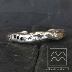 May include: A silver cuff bracelet with a textured surface and three small blue gemstones.