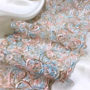 May include: Close-up of a delicate lace trim in shades of peach, light blue, and tan. The intricate floral and swirl patterns are visible. The lace is draped over a white satin fabric, creating a soft, elegant look.