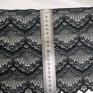 May include: Black lace fabric with a scalloped edge and a fan-like pattern. A metal ruler is placed vertically, showing measurements in centimetres. The fabric's intricate design is visible against a white background.