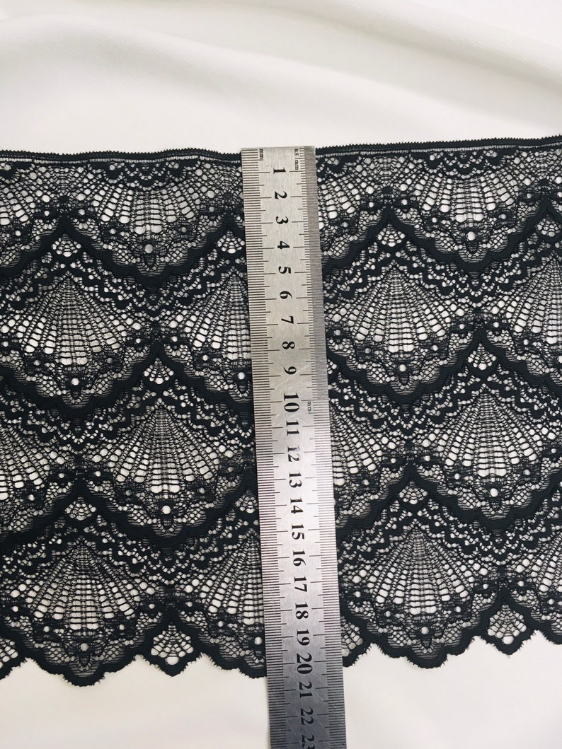 May include: Black lace fabric with a scalloped edge and a fan-like pattern. A metal ruler is placed vertically, showing measurements in centimetres. The fabric's intricate design is visible against a white background.