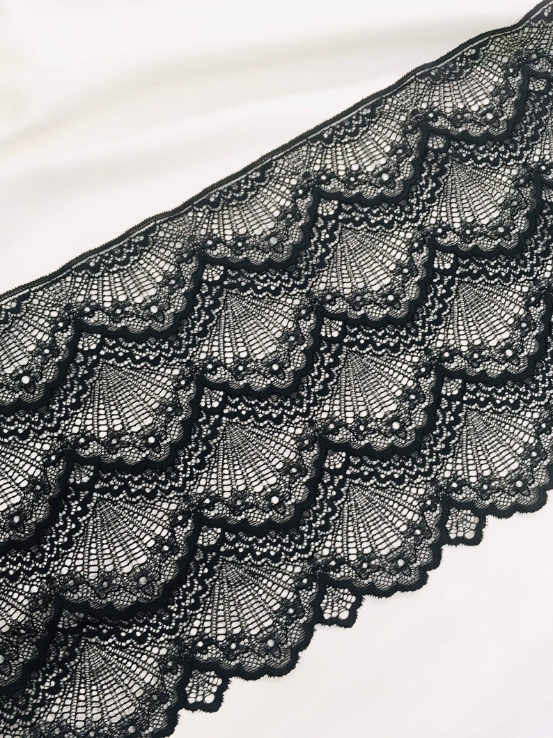 May include: Black lace trim with a scalloped edge. The lace has a repeating fan-like pattern with delicate detailing. The trim is set against a white background, highlighting the intricate design and texture of the lace.