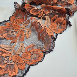 May include: Close-up of a floral lace trim. The trim features orange and tan flowers with black outlines and a black scalloped edge. The lace is set against a white background.