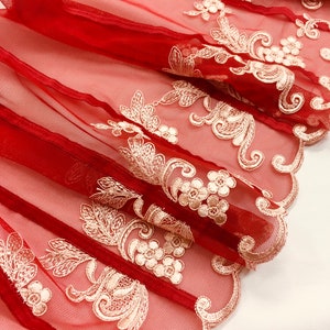 May include: Close-up of folded, sheer red fabric with cream-colored floral embroidery. The fabric has a scalloped edge and is layered, creating depth and texture. Suitable for crafting or decorative use.