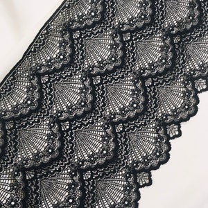 May include: Close-up of black lace fabric with a repeating geometric pattern. The lace features a scalloped edge and a delicate, open weave design. The fabric is set against a white background, highlighting the intricate details of the lace.