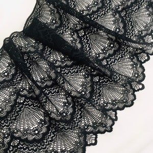 May include: Close-up of black lace fabric with a scalloped edge. The lace features a detailed, openwork design with a fan-like pattern. The fabric is folded, creating shadows and highlighting the intricate details of the lace.