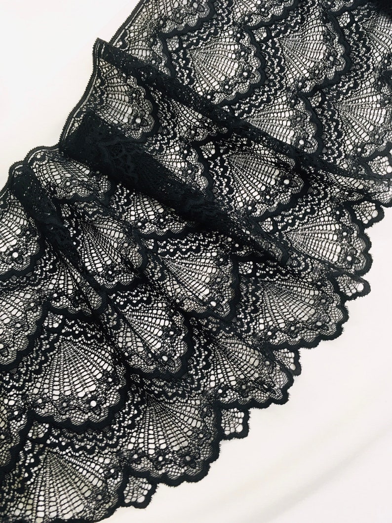 May include: Close-up of black lace fabric with a scalloped edge. The lace features a detailed, openwork design with a fan-like pattern. The fabric is folded, creating shadows and highlighting the intricate details of the lace.