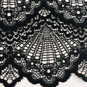 May include: Close-up of black lace fabric with a scalloped edge. The lace features an intricate, openwork design with a repeating pattern of floral and geometric shapes. The fabric is set against a white background.