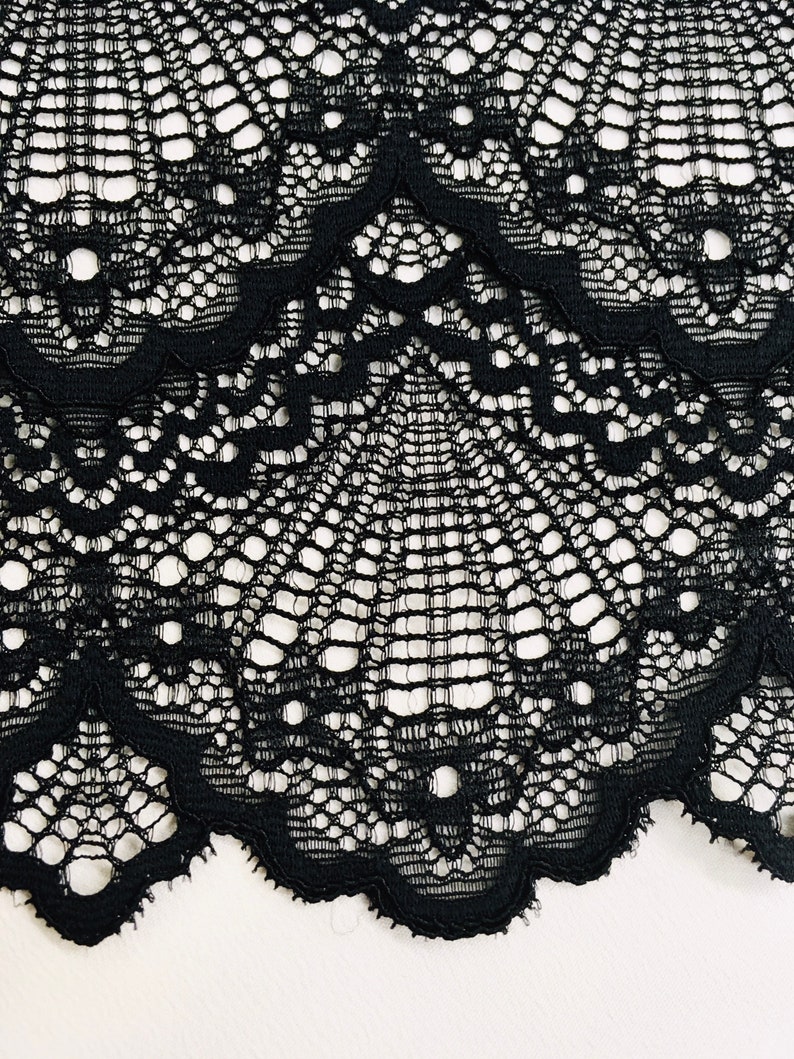 May include: Close-up of black lace fabric with a scalloped edge. The lace features an intricate, openwork design with a repeating pattern of floral and geometric shapes. The fabric is set against a white background.