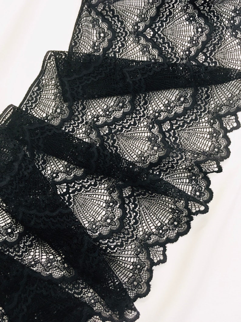 May include: Close-up of black lace fabric with a detailed, openwork design. The lace is folded, revealing layers of the intricate pattern. The fabric's delicate texture and dark colour create a striking visual contrast against the white background.