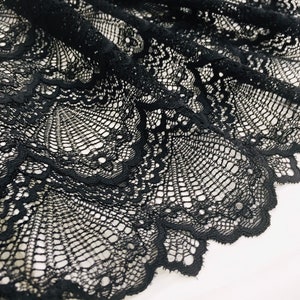 May include: Close-up of black lace fabric with an intricate, openwork design. The lace is draped, showing detailed patterns and textures. The fabric's scalloped edge adds a decorative touch. The lace is likely used for clothing or craft projects.