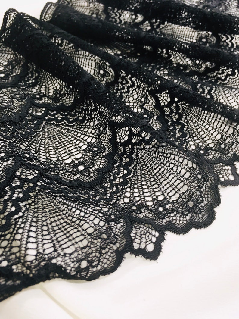 May include: Close-up of black lace fabric with an intricate, openwork design. The lace is draped, showing detailed patterns and textures. The fabric's scalloped edge adds a decorative touch. The lace is likely used for clothing or craft projects.