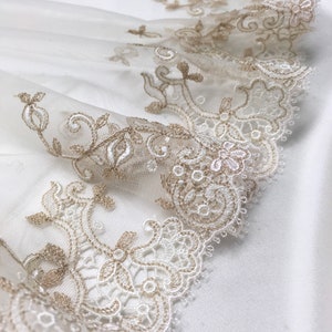 May include: Close-up of ivory lace fabric with detailed floral and scrollwork embroidery in a soft gold colour. The lace is layered over white fabric, creating a textured and elegant design. Suitable for bridal or craft projects.