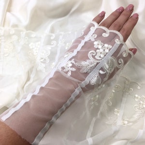 May include: Ivory bridal gloves with detailed floral lace. The gloves are made of sheer fabric, with a scalloped edge and small, sparkling embellishments. The design includes a decorative panel on the back of the hand. The gloves are a delicate accessory.