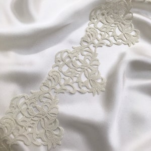 May include: A strip of off-white floral lace trim featuring an intricate, scalloped edge design with detailed embroidery. The lace is layered over a smooth, draped off-white satin fabric, creating a soft contrast in texture and material.