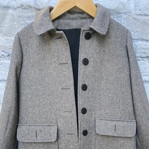 May include: A gray herringbone pattern coat with a black interior, two front pockets, and four dark buttons. The coat is hanging on a wooden hanger against a white brick wall.