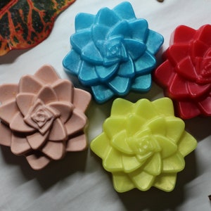 Op de afbeelding: Vier handgemaakte zeepjes in de vorm van bloemen. De zeepjes zijn in de kleuren blauw, roze, rood en geel.