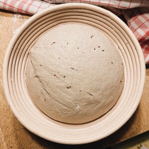 May include: A round loaf of bread dough rising in a  round, woven,  basket with a light brown color.