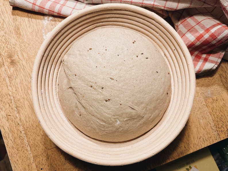 May include: A round loaf of bread dough rising in a  round, woven,  basket with a light brown color.