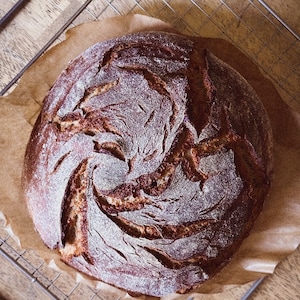 May include: A freshly baked loaf of sourdough bread with a dark brown crust and a swirl pattern on the top. The bread is sitting on a wire rack and parchment paper.