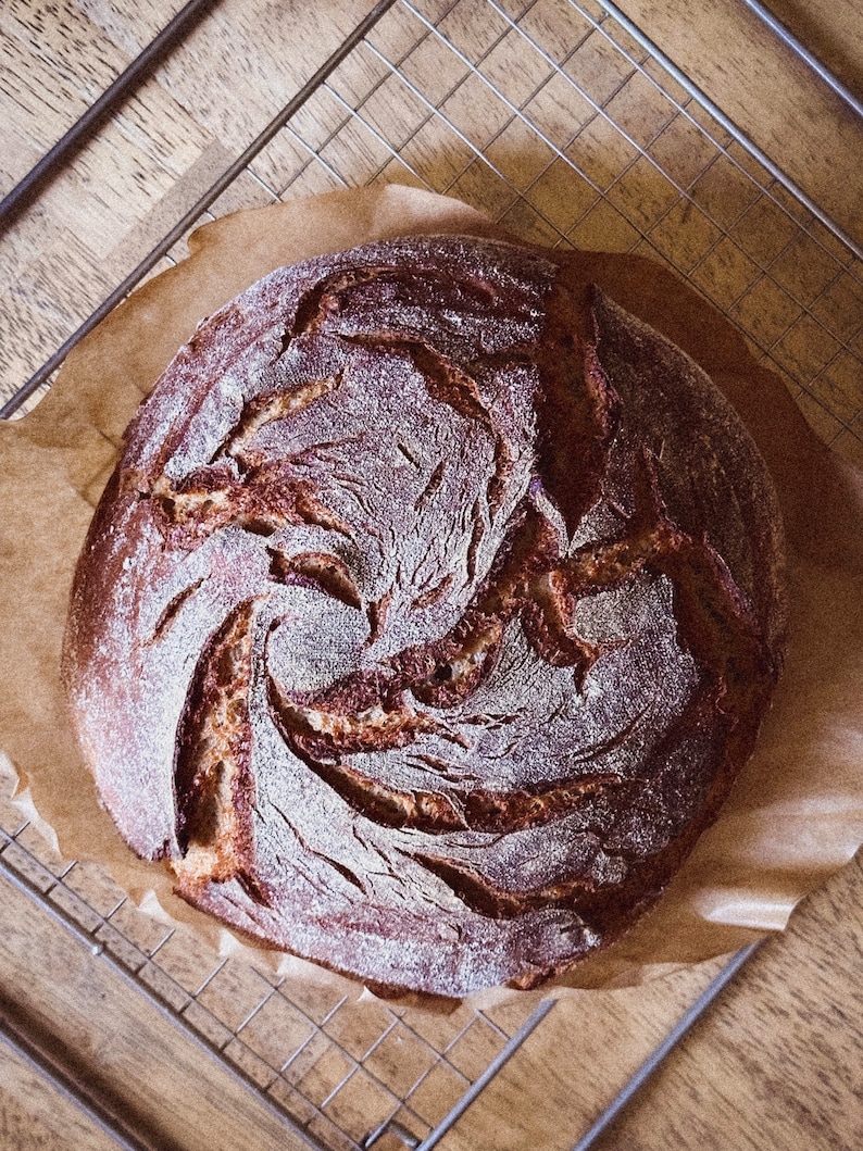 May include: A freshly baked loaf of sourdough bread with a dark brown crust and a swirl pattern on the top. The bread is sitting on a wire rack and parchment paper.