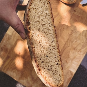 May include: A close-up of a sliced loaf of sourdough bread. The bread has a light brown crust and a soft, airy crumb. The bread is being held up by a hand, and the background is a wooden cutting board.