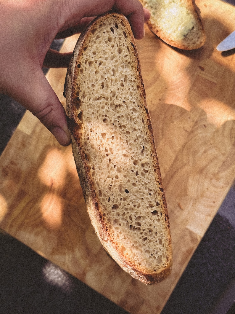 May include: A close-up of a sliced loaf of sourdough bread. The bread has a light brown crust and a soft, airy crumb. The bread is being held up by a hand, and the background is a wooden cutting board.