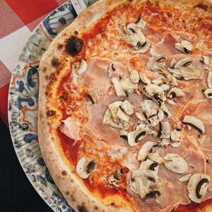May include: A close-up of a pizza with ham and mushrooms on a white and blue patterned plate. The pizza has a crispy crust and a red sauce.
