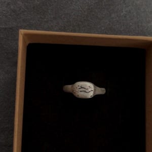 Puede incluir: Un pequeño anillo plateado con una cara rectangular con un diseño ondulado. El anillo se presenta en una caja de cartón marrón con interior negro, sobre un fondo gris.