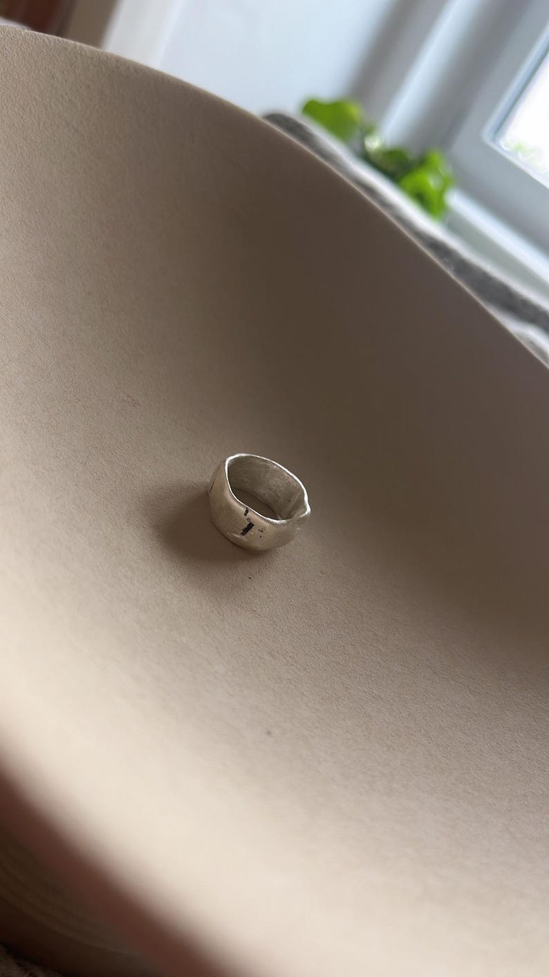 May include: A silver ring with a textured, irregular shape. The ring has a small, vertical line etched into the surface.