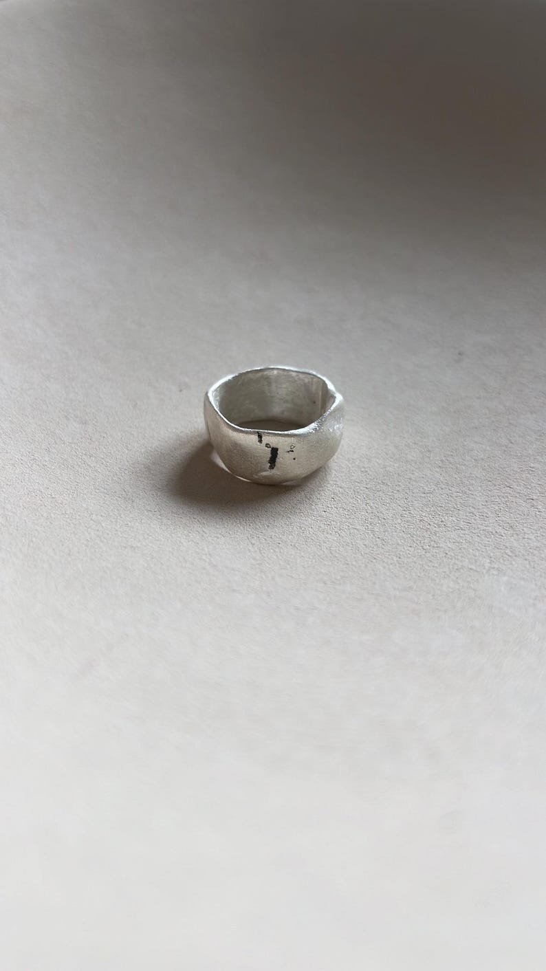 May include: A silver ring with a textured, irregular shape. The ring has a slightly hammered finish.