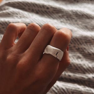 May include: A silver ring with a textured, slightly curved surface. The ring is worn on a finger.