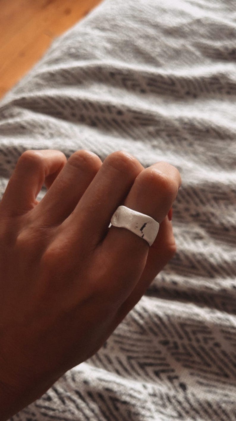 May include: A silver ring with a textured, slightly curved surface. The ring is worn on a finger.