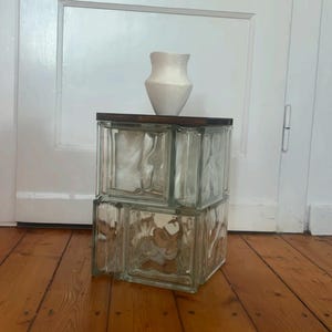 Puede incluir: Una mesa auxiliar de bloques de vidrio transparente con una tapa de madera oscura, que sostiene un jarrón de cerámica blanca. La mesa está construida con dos bloques de vidrio apilados, creando una estética moderna única.