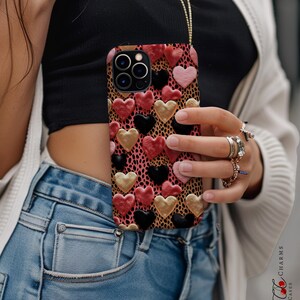 May include: A phone case with a pink, black, and gold heart pattern on a leopard print background. The case is being held by a person wearing blue jeans.