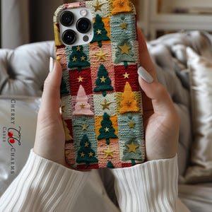 Op de afbeelding: Een telefoonhoesje met een kerstboompatroon. Het hoesje is gemaakt van gebreid materiaal in verschillende kleuren, waaronder rood, groen, blauw en geel. Het hoesje heeft een herhalend patroon van kerstbomen en sterren.