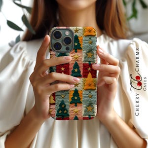 Op de afbeelding: Een telefoonhoesje met een kerstthema gebreid patroon met groene bomen, gouden sterren en rode en witte strepen. De hoes is gemaakt voor een telefoon met een drievoudige lenscamera.