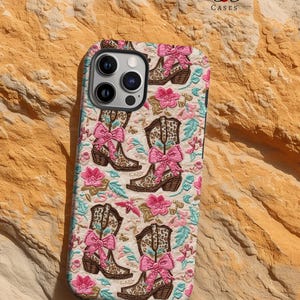 May include: A white phone case featuring a pattern of brown cowboy boots with pink bows and floral accents. The case has a cutout for the camera lens and the text "Cherry Charms Cases" at the top.
