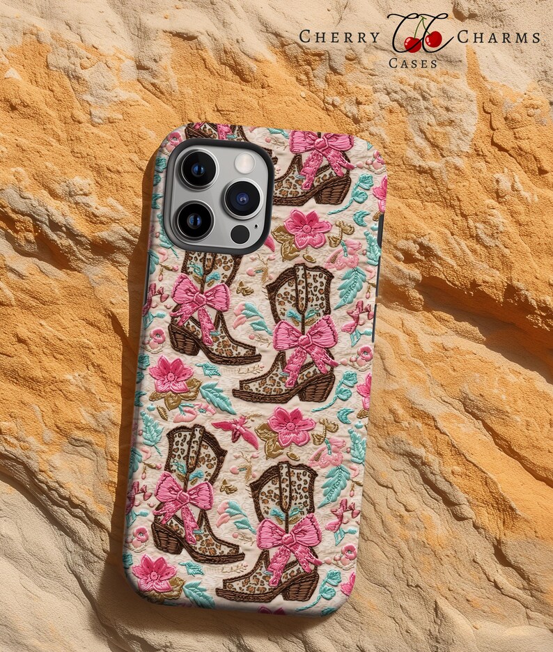 May include: A white phone case featuring a pattern of brown cowboy boots with pink bows and floral accents. The case has a cutout for the camera lens and the text "Cherry Charms Cases" at the top.