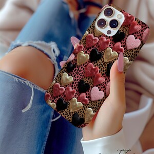 May include: A phone case with a pink, black, and gold heart pattern on a leopard print background. The case has a raised lip around the camera lens.