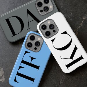 Op de afbeelding: Drie telefoonhoesjes met verschillende achtergrondkleuren en zwarte letters. Het eerste hoesje is grijs met de letter "D", het tweede hoesje is wit met de letters "KC" en het derde hoesje is blauw met de letters "TF".