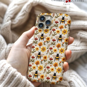 Puede incluir: Una funda blanca para iPhone con un patrón de abejas y girasoles bordados. La funda tiene un recorte para la cámara y un logotipo que dice "Cherry Charms Cases".