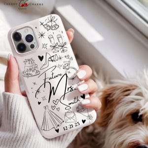 May include: A white phone case with a black line art design featuring wedding-themed illustrations, including a cake, dress, and ring. The text "Future Mrs. Smith" and the date "12-21-25" are also displayed.