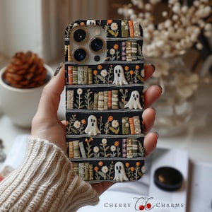 Pode incluir: Capa de telefone preta com um design de estante com figuras de fantasmas brancos, flores laranja e brancas e várias lombadas de livros. A capa tem uma aparência texturizada e o texto "CHERRY CHARMS CASES" é visível.