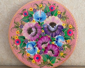 May include: A pink round wooden plate with a hand-painted floral design. The flowers are in shades of pink, purple, blue, and yellow. The plate has a gold trim around the edge.