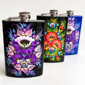 May include: Three metal flasks with screw-top caps, each featuring hand-painted floral designs. The front flask is black with purple and light blue flowers. The middle flask is black with red and blue flowers. The back flask is blue with purple flowers.