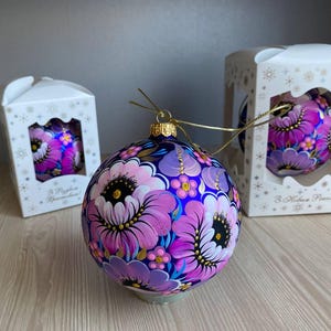 Puede incluir: Tres adornos de vidrio pintados a mano con diseños florales. Los adornos son morados con flores rosas y blancas y detalles dorados. Los adornos están en cajas blancas con el texto "3 Новые Розы!" en la parte delantera.