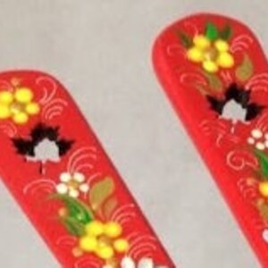 May include: Two red wooden spoons with floral designs. Each spoon features yellow, white, and black flowers with green leaves and gold accents. The spoons have a curved handle and a flat, rectangular head.