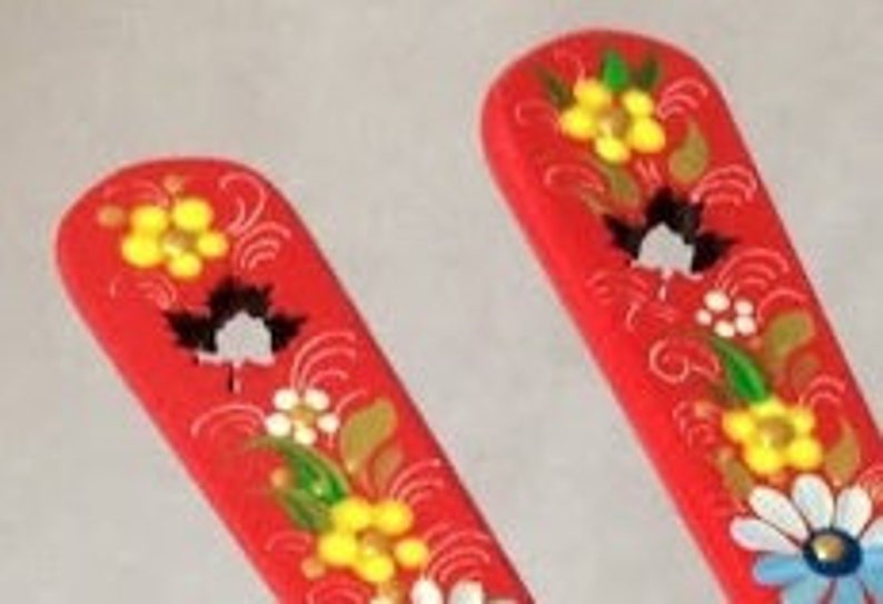 May include: Two red wooden spoons with floral designs. Each spoon features yellow, white, and black flowers with green leaves and gold accents. The spoons have a curved handle and a flat, rectangular head.