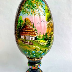 Puede incluir: Un huevo de madera pintado a mano con una escena de una pequeña casa en un bosque. El huevo está sobre un soporte negro con detalles dorados y está decorado con flores y hojas.